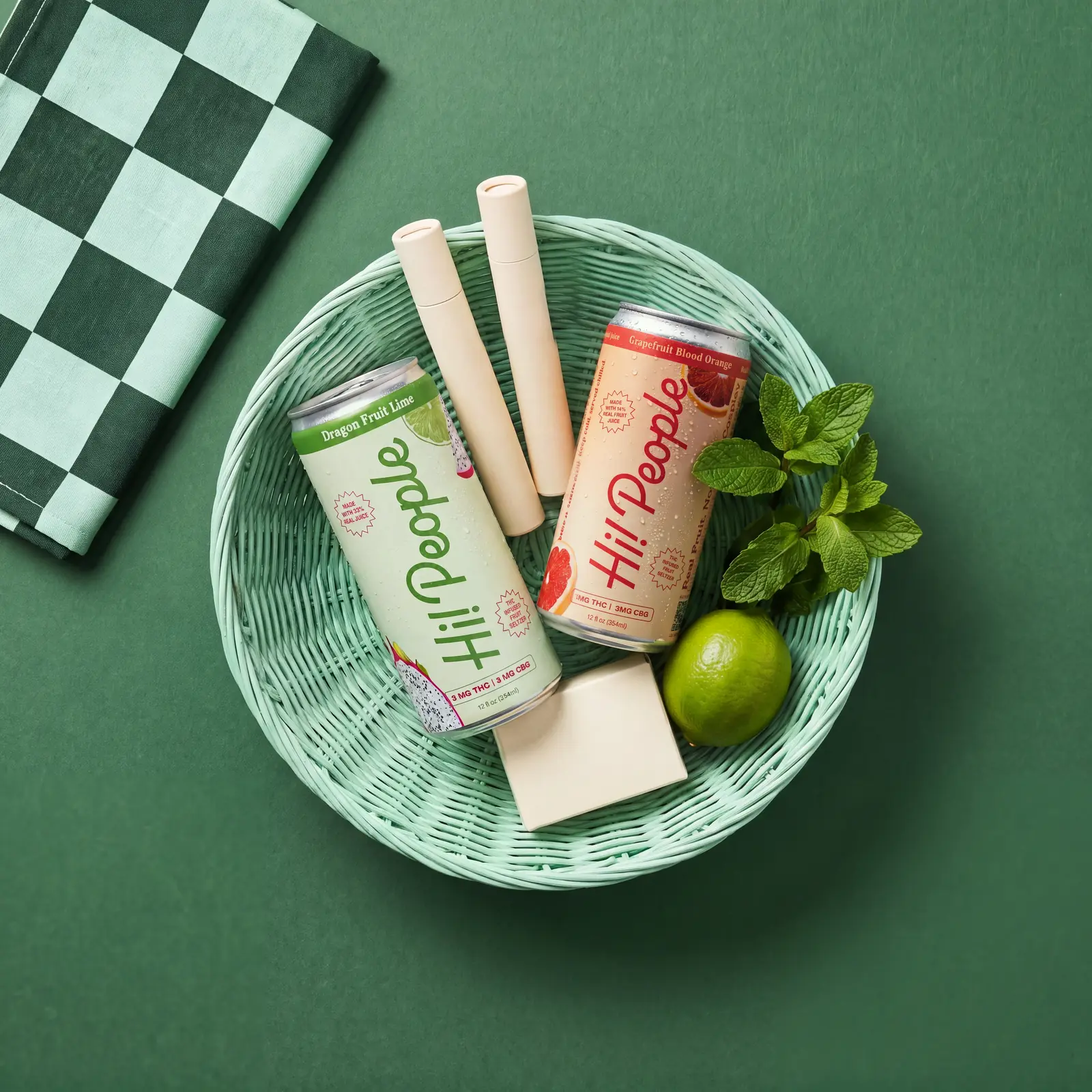 A mint market basket holding Hi! People Grapefruit Blood Orange and Dragon Fruit Lime seltzer cans, two pre-roll tubes, fresh mint and a lime
